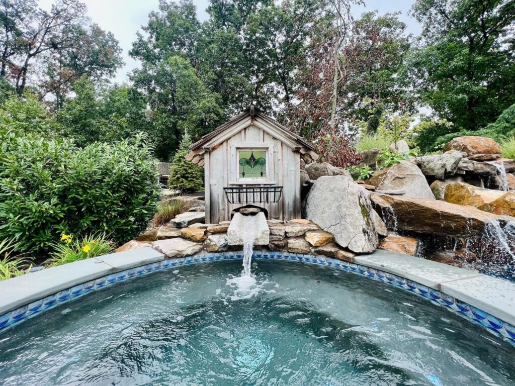 A fountain pours into a custom pool surrounded by lush trees and a rock formation.